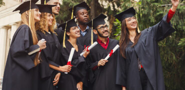 Group of multiethnic students taking selfie on smartphone to celebrate their graduation. Education, qualification and gown concept.