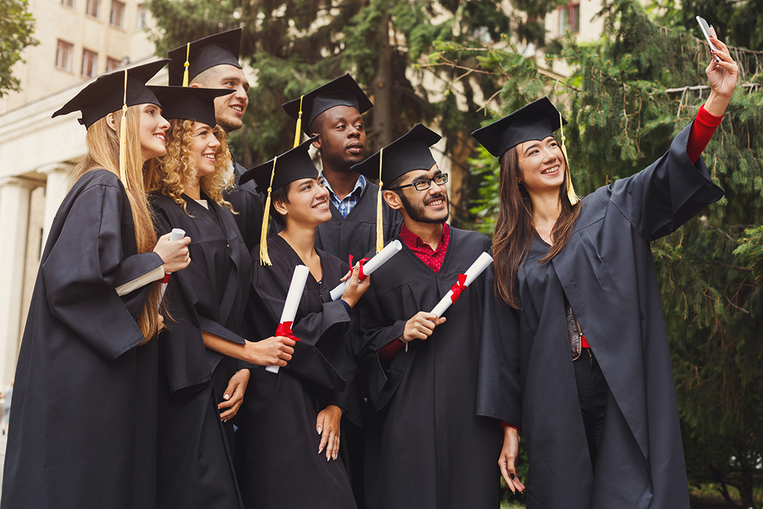 Group of multiethnic students taking selfie on smartphone to celebrate their graduation. Education, qualification and gown concept.