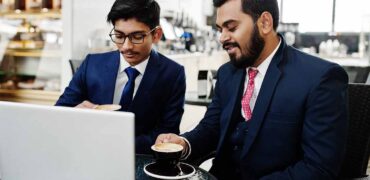 Two indian business man in suits sitting at office on cafe, looking at laptop and drinking coffee.