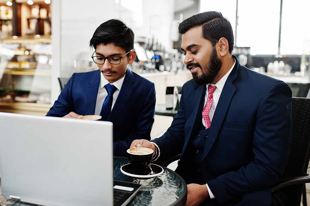 Two indian business man in suits sitting at office on cafe, looking at laptop and drinking coffee.